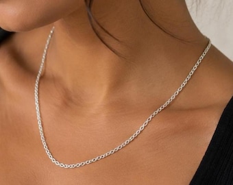 May include: A simple silver-toned chain necklace featuring small, uniform oval-shaped links. The metallic chain rests against the neck, showcasing a polished finish and a classic, continuous design that connects around the collarbone area.