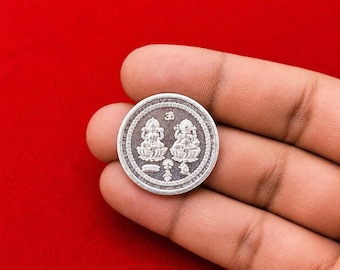 Könnte beinhalten: Eine kreisförmige silberfarbene Münze mit geprägten Figuren von Lakshmi und Ganesha, die auf Lotusblumen sitzen. In der oberen Mitte befindet sich das Om-Symbol, während der untere Teil kleine dekorative Elemente aufweist.