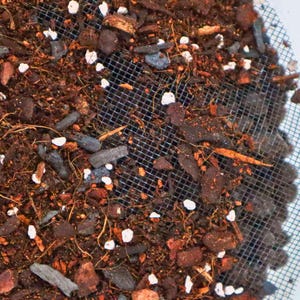 May include: Close-up of a brown soil mixture with white perlite and small pieces of bark. The soil is in a mesh container.