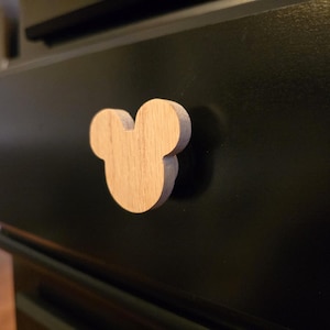 May include: A wooden Mickey Mouse-shaped cabinet knob. The knob is light brown and mounted on a black cabinet drawer. The knob is a decorative accent for furniture.