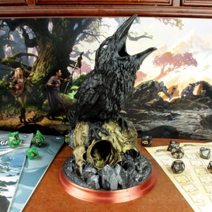 Könnte beinhalten: Eine schwarze Rabenfigur mit offenem Schnabel, die auf einem schädelförmigen Sockel sitzt. Die Figur ist von grünen Würfeln und einem Spielbrett mit dem Text "Der Drache von Eisnadelgl" umgeben.