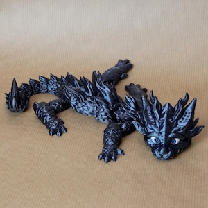 Könnte beinhalten: Eine schwarze Drachenfigur mit blauen Augen, die auf einer braunen Oberfläche liegt. Der Drache hat einen stacheligen Rücken und Schwanz.