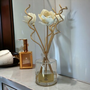 May include: A clear glass diffuser bottle with a gold label that reads "Rose Garden". The bottle is filled with a clear liquid and has wooden sticks inserted into it. The sticks are topped with white ceramic flowers.