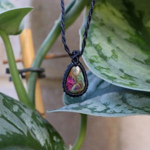 May include: A teardrop-shaped pendant featuring a multicoloured stone with prominent shades of magenta, green, and gold, encased in a black macrame cord setting. The pendant hangs from a matching black braided cord necklace.