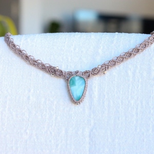 May include: A macrame necklace featuring a teardrop-shaped light blue stone pendant set in a tan woven cord. The cord has an intricate braided pattern with small metallic beads integrated into the design, creating a detailed and textured appearance.