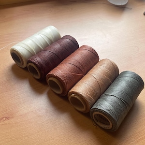 Pode incluir: Seis carretéis de linha em várias cores, incluindo creme, bordô e tons de castanho e cinza. Os carretéis estão dispostos diagonalmente sobre uma superfície de madeira, mostrando os diferentes tons da linha.