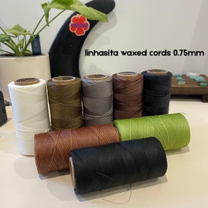 May include: A selection of waxed cord spools in shades of white, brown, grey, and black. The text reads "linhasita waxed cords 0.75mm". The cords are tightly wound on cardboard spools, ideal for crafts and jewellery making.