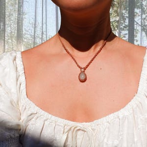 May include: A necklace featuring a braided brown cord chain with a small white pearl accent and a central oval-shaped light green gemstone pendant. The necklace is worn around the neck, resting against a white blouse with puffed sleeves.