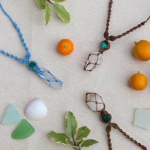May include: Overhead shot of three macrame necklaces with crystal pendants, in blue and brown. The necklaces have green beads. The image is styled with seashells, small oranges, and greenery on a neutral background.
