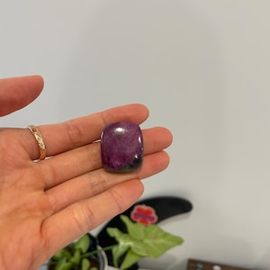 May include: A polished, rectangular gemstone with a deep magenta hue, accented by areas of green and black. The stone is held in a hand, showcasing its smooth, rounded edges and natural colour variations. The gemstone is approximately 3.8 cm long.