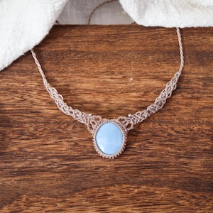 May include: A macrame necklace with a pale blue oval pendant. The necklace is displayed on a wooden surface with a white cloth and two crystals, one clear and one purple.
