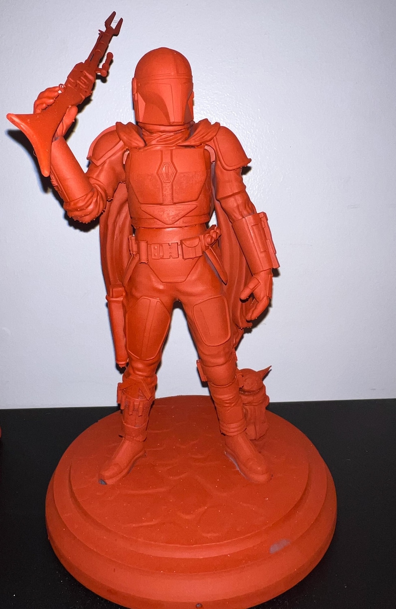 Pu&ograve; includere: Una statuetta dipinta di rosso di un personaggio di Star Wars, The Mandalorian, che tiene un fucile blaster. The Mandalorian &egrave; in piedi su una base rotonda con una piccola creatura, Grogu, ai suoi piedi.
