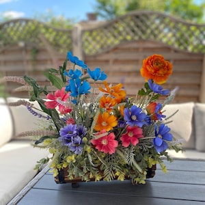 May include: A colourful floral display in a wooden planter. The arrangement includes blue, orange, pink, and purple flowers, with green foliage. The rectangular planter sits on a dark surface. The flowers are a mix of sizes.