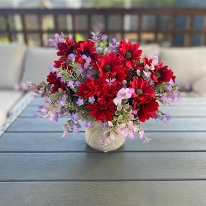 May include: A floral display with striking red flowers and purple accents, set in a textured vase. The arrangement includes green foliage and is placed on a grey wooden table. The flowers are a focal point, offering a burst of colour and texture.