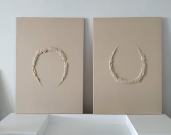 Op de afbeelding: Twee beige canvas schilderijen met verhoogde, getextureerde cirkels in het midden. De cirkels zijn onvolledig en lijken op een halve maan.