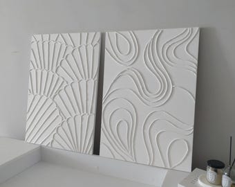 May include: Two white canvas wall art pieces with raised 3D designs. One piece features a geometric pattern of overlapping circles and the other piece features a pattern of overlapping fan shapes.