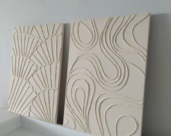 Op de afbeelding: Twee off-white canvas schilderijen met reliëf textuurontwerpen. Een schilderij heeft een waaiervormig patroon en de andere heeft een golvend, abstract patroon.