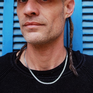 May include: A man with dreadlocks wearing a black shirt and a dark green baseball cap with white text. He is standing in front of a blue wooden shutter.