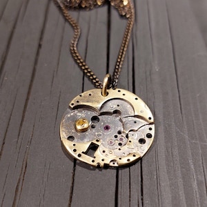 Może przedstawiać: Złoty naszyjnik z zawieszką wykonaną z vintage'owego cyferblatu zegarka. Zawieszka ma okrągły kształt i widoczne są na niej różne koła zębate i zębatki. Naszyjnik leży na ciemnym drewnianym tle.