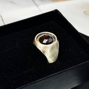 May include: A silver ring with a dark red gemstone set in a gold bezel. The ring is displayed in a black velvet box.