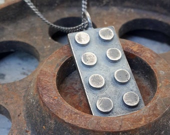 Könnte beinhalten: Eine rechteckige Silber-Anhänger-Halskette mit einem Lego-Stein-Design. Der Anhänger hat sechs erhabene kreisförmige Noppen und hängt an einer Silberkette. Der Anhänger ist vor einem rostigen, industriellen Hintergrund platziert.