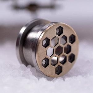 May include: A silver and gold ear gauge with a hexagonal pattern. The gauge is made of metal and has a smooth finish.
