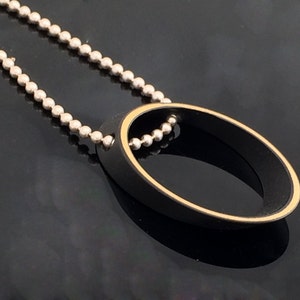 Op de afbeelding: Een zwarte ovale hanger met een gouden rand hangt aan een zilveren ketting. De hanger is een eenvoudig, modern ontwerp.