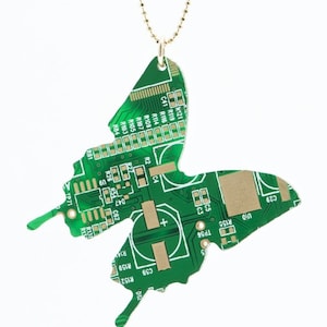 May include: A green circuit board shaped like a butterfly, with gold-colored components, hangs from a gold chain. The circuit board has text and numbers printed on it, including "R119", "R107", "R105", "R103", "R26", "R54", "C4", "R2", "C62", "C29", "U10", "R155", "TP56", "R159", "R152", "C59", "R141", "R116", "R114", "R109", "C41", "K121", "TP71", "R131", "R13", "012", and "33".