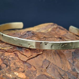 Peut inclure: Un bracelet manchette de couleur or avec une surface texturée et des marques gravées. Le bracelet ouvert est présenté sur une surface de pierre rugueuse, marron et orange, sur un fond bleu foncé.