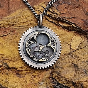 Może przedstawiać: Srebrny naszyjnik z okrągłym wisiorem, który prezentuje szczegółowy wzór koła zębatego z złotymi akcentami. Wisiorek wykonany jest z ponownie wykorzystanych części zegarków.