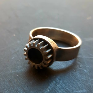 May include: A silver ring with a gear design. The gear is centered on the ring and has a circular opening in the middle.