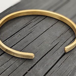 Peut inclure: Un bracelet jonc simple en métal doré. Le bracelet est ouvert et a un profil plat légèrement arrondi.