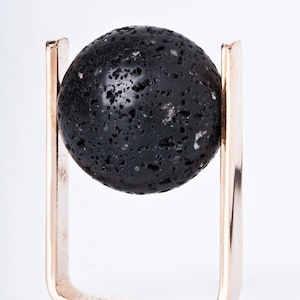 Könnte beinhalten: Eine schwarze Lavasteinkugel sitzt auf einem goldenen Metallständer. Der Ständer ist einfach rechteckig mit abgerundeten Kanten.