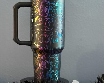 May include: A black stainless steel travel mug with a handle and a rainbow iridescent finish. The mug is decorated with a Halloween-themed pattern of ghosts, pumpkins, bats, and stars. The mug has a straw and is sitting on a black and white garland.