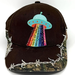 Puede incluir: Gorra de béisbol marrón con visera de camuflaje y un parche decorativo de un OVNI azul claro que emite un rayo arcoíris. La gorra tiene una visera curva y un diseño clásico.