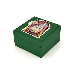 May include: A square, dark green wooden box with a decorative sticker on the lid. The sticker features a vintage illustration of a woman in a flowing dress. The box is likely used for storage or display.