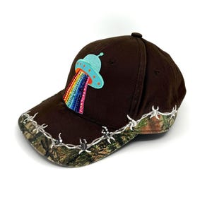 Może przedstawiać: Brązowa czapka bejsbolowa z kamuflażowym daszkiem i białym wzorem z drutu kolczastego. Czapka posiada naszywkę z jasnoniebieskim UFO emitującym tęczową wiązkę. Unikalny i przyciągający wzrok dodatek.