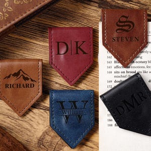 Może przedstawiać: Kolekcja spersonalizowanych skórzanych zakładek do książek w różnych kolorach, w tym brązowym, bordowym i niebieskim. Każda zakładka zawiera spersonalizowany grawerowany tekst, taki jak inicjały, imiona i grafika gór. Ułożone na drewnianej powierzchni.