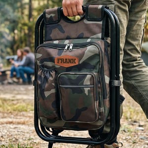 May include: A camouflage print backpack chair with a black metal frame, featuring a brown leather-like patch with the name FRANK embossed on the front, a side pocket, a front zippered compartment, and a top handle for easy carrying.