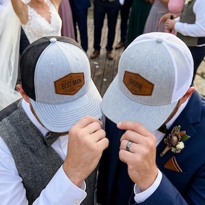Peut inclure: Deux casquettes de baseball avec des dos en filet et des panneaux avant gris clair. Une casquette présente un écusson en similicuir marron avec le texte BEST MAN, l'autre avec le texte GROOM. Les deux casquettes ont une visière structurée.