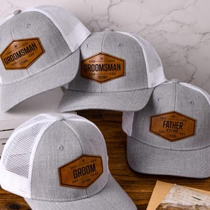 Peut inclure: Quatre casquettes trucker grises et blanches avec des écussons en similicuir marron. Les écussons hexagonaux portent les inscriptions GROOMSMAN, FATHER OF THE BRIDE ou GROOM, accompagnées de la date 7.21.2026 et de motifs de flèches.