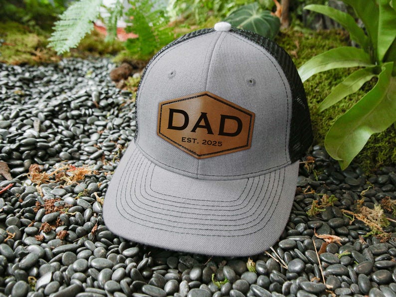 May include: A gray trucker hat with a black mesh back and a brown leather patch. The patch features the word "DAD" and "EST. 2025" in black lettering. The hat is resting on a bed of black pebbles.