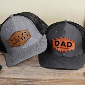 May include: Two baseball caps with mesh backs and leather patches. One is gray with a black mesh back and a patch that says "DAD" with names. The other is dark gray with a black mesh back and a patch that says "DAD" with names.