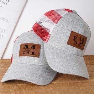 Puede incluir: Dos gorras de béisbol grises con parches de cuero. Un parche dice "Martin" encima de "IVI", el otro tiene "AMADEO" con un diseño de cornamenta de ciervo. Una gorra tiene la parte trasera de malla a cuadros rojos y blancos.