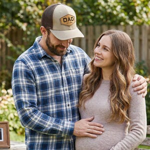 May include: A couple stands outdoors, the man wearing a baseball cap with "DAD" on it. The woman is pregnant. A framed ultrasound image sits on a wooden bench. The man wears a blue plaid shirt.