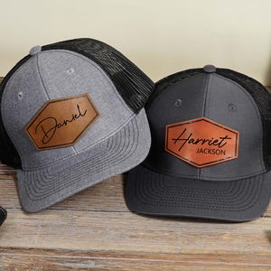 May include: Two personalised trucker caps. One is grey with a black mesh back and a brown leather patch that says "Daniel". The other is dark grey with a black mesh back and a brown leather patch that says "Harriet Jackson".