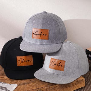 Op de afbeelding: Drie baseballpetten in zwart en grijs, elk met een bruin leren patch. De patches zijn voorzien van de namen "Andrew", "Oliver" en "Carter" in een cursief lettertype. De petten worden op een houten oppervlak weergegeven.