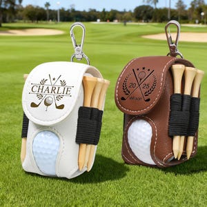K&ouml;nnte beinhalten: Zwei Golfballtaschen, eine wei&szlig;e und eine braune, jeweils mit einem Golfball, Tees und einem silbernen Karabinerhaken. Auf der wei&szlig;en Tasche steht der Name "Charlie". Auf der braunen Tasche stehen die Zahlen "20 25".