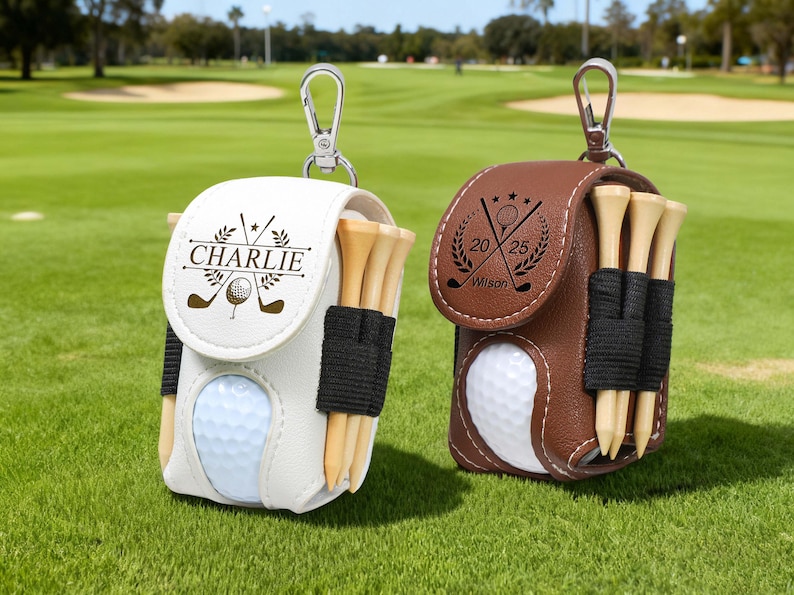 K&ouml;nnte beinhalten: Zwei Golfballtaschen, eine wei&szlig;e und eine braune, jeweils mit einem Golfball, Tees und einem silbernen Karabinerhaken. Auf der wei&szlig;en Tasche steht der Name "Charlie". Auf der braunen Tasche stehen die Zahlen "20 25".