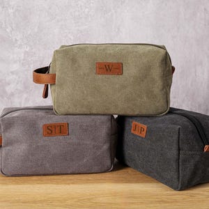 Könnte beinhalten: Drei Kulturbeutel aus Canvas mit Lederakzenten und personalisierten Initialen. Die obere Tasche ist grün mit den Initialen "W", die mittlere Tasche ist grau mit den Initialen "SIT" und die untere Tasche ist schwarz mit den Initialen "JP".